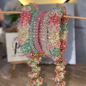 May include: A set of multi-coloured beaded bangles featuring faceted crystals in shades of pink, teal, and clear. The bangles are adorned with small, translucent floral accents and dangling beaded strands, all connected by thin gold-toned wire.