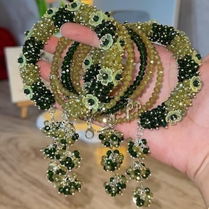 May include: A set of circular bracelets crafted from alternating dark green and light olive green faceted beads. The bracelets feature small, pale green fabric flower accents and dangling clusters of dark green beads attached with silver-toned metal links.