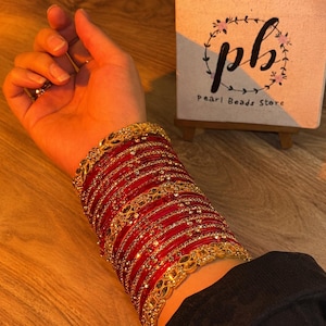 Pode incluir: Uma pilha de pulseiras vermelhas e douradas com padrões metálicos intrincados e pequenas pedras transparentes incrustadas. O conjunto inclui várias faixas vermelhas finas intercaladas com pulseiras de metal dourado mais largas e ornamentadas.