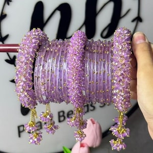 Op de afbeelding: Een set lavendelkleurige armbanden bestaande uit een mix van getextureerde, doorschijnende paarse banden en kralenbanden. De armbanden met kralen zijn versierd met kleine goudkleurige accenten en bungelende trosjes lavendelkleurige kralen.