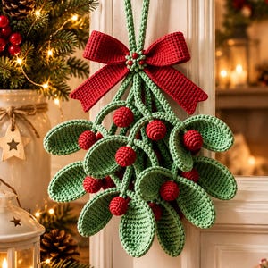 Puede incluir: Una decoración de muérdago tejida a ganchillo con hojas verde salvia y bayas de color rojo brillante, unidas por un gran lazo rojo tejido. El adorno cuelga de un lazo de ganchillo verde, mostrando texturas de hilo y un diseño festivo.