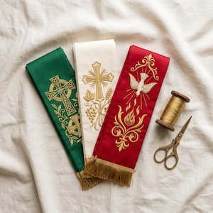 May include: Three liturgical stoles in green, white, and red, each featuring intricate gold embroidery of a Celtic cross, a Latin cross with wheat and grapes, and a dove with flames. A wooden spool of gold thread and metal scissors are included.