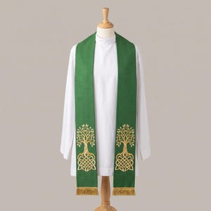 May include: A green liturgical stole featuring gold embroidered tree designs with intricate knotwork roots at the base of each panel. The stole is draped over a white alb and includes gold fringe trim at the bottom edges of both vertical panels.