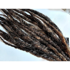 May include: A collection of synthetic hair extensions styled as dreadlocks, featuring a blended color palette of dark brown and lighter golden brown tones. The individual locks have a textured, matted appearance with loose fibers throughout the strands.