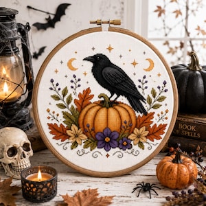 May include: A circular wooden embroidery hoop containing a cross-stitch design of a black raven perched on an orange pumpkin, surrounded by autumn leaves, purple flowers, and small stars and crescent moons on a white fabric background.