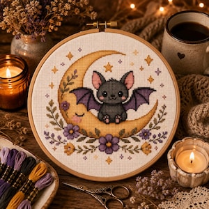 Può includere: Un telaio da ricamo circolare in legno contenente un motivo a punto croce di un piccolo pipistrello grigio con ali viola seduto su una luna crescente gialla, decorato con fiori viola, foglie verdi e piccole stelle gialle su tessuto bianco.