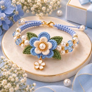 May include: A bracelet featuring a central crocheted blue and cream flower with green leaves, accented by white pearls, gold beads, and light blue crystals. The band is made of braided blue cord with a gold lobster clasp and a small white flower charm.