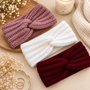 May include: Three knitted headbands featuring a twisted front design. One headband is mauve, one is white, and one is deep burgundy. Each piece displays a consistent ribbed texture throughout the fabric, showcasing a simple and functional construction.