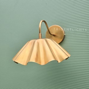 May include: A brass wall sconce featuring a curved arm and a wide, scalloped, bell-shaped shade. The circular mounting plate is attached to the wall, and the word TRIDENTLIGHTS is printed in white text on the right side of the fixture.