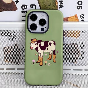 May include: A sage green smartphone case featuring a graphic illustration of a white and brown calf wearing a tan cowboy hat and matching tan cowboy boots, standing among small pink, yellow, and white flowers on the back of the case.