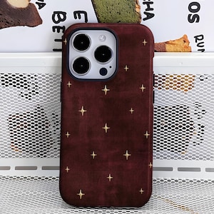 May include: A deep burgundy smartphone case featuring a repeating pattern of small, four-pointed gold stars. The case has a cutout for a triple-lens camera system on the upper left side and a smooth, matte-textured finish across the back surface.
