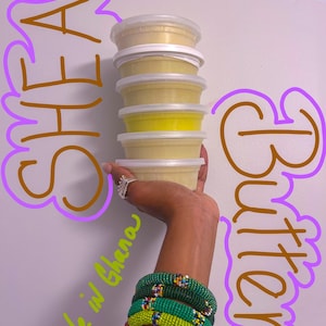 Puede incluir: Una mano sosteniendo una pila de cuatro recipientes de manteca de karité, etiquetados como "Shea Butter" y "Made in Ghana". Los recipientes son de plástico transparente con tapas blancas. La mano lleva varias pulseras de cuentas verdes.