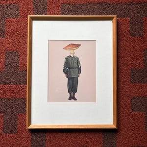 Puede incluir: Impresión artística enmarcada que presenta a una persona con uniforme militar y un sombrero de seta. La obra de arte está sobre un fondo rosa pálido y enmarcada en un marco de madera natural. El fondo es una alfombra estampada en rojo y marrón.