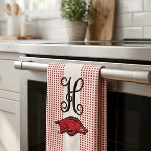 May include: A white kitchen towel with a red checkered pattern, featuring a large black embroidered letter H and a red razorback hog mascot graphic. The towel is finished with three red fabric tassels hanging from the bottom edge.