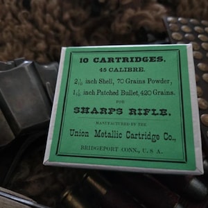 May include: A rectangular box of 10 cartridges for a Sharps rifle, featuring a green label with black text that reads 10 CARTRIDGES, 45 CALIBRE, 2 1/10 inch Shell, 70 Grains Powder, 1 1/10 inch Patched Bullet, 420 Grains, Union Metallic Cartridge Co.