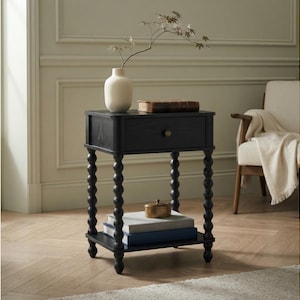 May include: A black wooden nightstand featuring a single drawer with a small round knob, four spindle-style legs with a bobbin design, and a lower shelf. The top surface holds a cream-colored vase and a book, while the shelf holds two books.