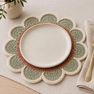 Op de afbeelding: Een rond, bloemvormig gehaakt placemat met een centrale ring van terracotta garen, omringd door saliegroene bloemblaadjes en afgewerkt met een crèmekleurige rand. Een effen, gebroken wit keramieken bord staat in het midden van het placemat.