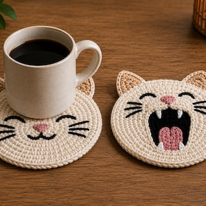 May include: Two round, cream-colored crochet coasters shaped like cat faces. One coaster features a sleeping cat with closed eyes and a pink nose, supporting a speckled ceramic mug filled with black coffee. The other shows a yawning cat.