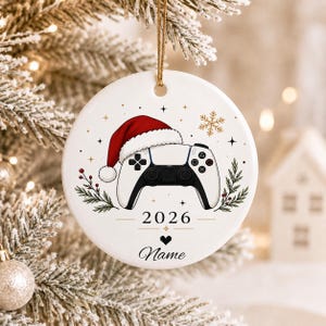 May include: A round white ceramic ornament featuring a black and white video game controller wearing a red Santa hat. The design includes a gold snowflake, small stars, green pine sprigs, the year 2026, a pixelated heart, and the text Name.