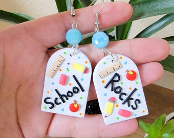 May include: White polymer clay earrings with a school theme. One earring says "School" and the other says "Rocks". Both earrings have a ruler, pencil, eraser, and apple design.