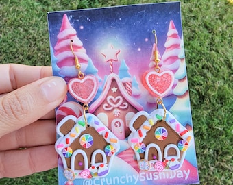 May include: A pair of gingerbread house earrings with pink heart charms. The earrings are made of polymer clay and have a white glaze. The gingerbread houses are decorated with colorful sprinkles and candy.