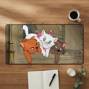 Könnte beinhalten: Eine rechteckige Schreibtischunterlage mit einer farbenfrohen Illustration von drei Kätzchen aus Aristocats. Das Design zeigt ein orangefarbenes Kätzchen, ein weißes Kätzchen mit einer rosa Schleife und ein braunes Kätzchen am Fenster.