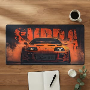 Puede incluir: Una alfombrilla de escritorio rectangular que presenta un coche deportivo Toyota Supra de color naranja vibrante centrado sobre un fondo oscuro con humo naranja y la palabra SUPRA en letras grandes, negritas y desgastadas.