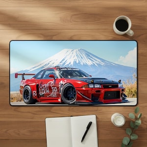 Puede incluir: Una alfombrilla de escritorio rectangular que muestra un coche deportivo rojo brillante con capó negro y calcomanías blancas estilo grafiti. El coche está aparcado frente a una montaña nevada bajo un cielo azul, capturando un vehículo.