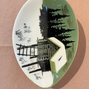 May include: An oval ceramic dish featuring a hand-painted scene of a log cabin nestled among black pine trees against a sage green background. The design includes a black ladder-like structure and the handwritten text "Mitt G. fur" in white.