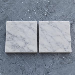 May include: Two square coasters made of white marble with natural gray veining. Each coaster features a smooth, flat surface and straight edges, showcasing the unique stone patterns characteristic of polished white marble material.