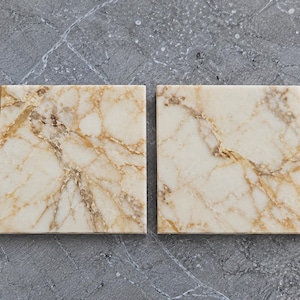 May include: Two square coasters made of cream-colored stone with prominent golden-brown veining throughout the surface. Each coaster features a smooth, polished finish and distinct natural patterns that vary slightly between the two pieces.