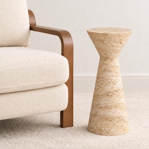 May include: A cylindrical side table crafted from beige, porous travertine stone. The table features a distinct hourglass silhouette with a wider circular top and base that taper inward toward the center, showcasing the natural texture of the stone.