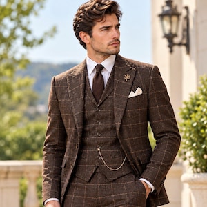 Pode incluir: Um fato de três peças aos quadrados castanhos composto por um blazer, um colete abotoado com uma corrente dourada e calças a condizer. O fato é usado com uma camisa branca de colarinho, uma gravata castanha texturada, um lenço e um alfinete.