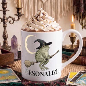 May include: A white ceramic mug filled with whipped cream topped with sprinkles. The mug features a graphic of a green frog wearing a black witch hat and riding a broomstick in front of a crescent moon, with the word PERSONALIZE printed below.