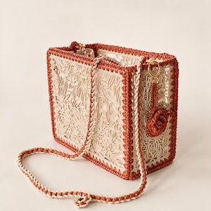 Può includere: Una borsa rettangolare con un motivo floreale color crema tagliato al laser sui lati e sulla base. La struttura è rinforzata con un bordo all'uncinetto color ruggine e una tracolla abbinata. Un piccolo ciondolo a forma di fiore all'uncinetto color ruggine pende da un anello dorato.