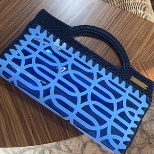 以下が含まれることがあります： 濃いネイビーブルーのクロシェ編みのトップとハンドルを備えた長方形のハンドバッグ。ライトブルーのレーザーカットされた幾何学模様のオーバーレイが施されています。オーバーレイは深いブルーの半透明のベースの上にあり、小さな長方形のゴールドトーンの金属タグが付いています。