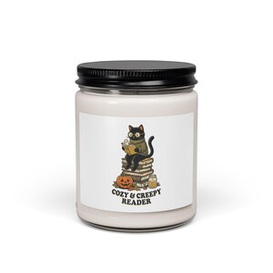 Pode incluir: Uma vela branca num frasco de vidro com tampa preta, com um rótulo que mostra um gato preto de óculos e camisola, a ler um livro sentado numa pilha de livros ao lado de uma abóbora e uma vela, com o texto COZY & CREEPY READER.