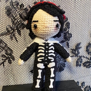 Könnte beinhalten: Eine gehäkelte Puppe mit schwarzem Haar und einem roten Stirnband, die ein schwarzes langärmeliges Outfit mit einem weißen Skelettknochen-Aufdruck auf der Vorderseite, einem weißen Kragen und hellen Händen trägt und aufrecht steht.