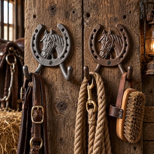May include: Two cast iron wall hooks shaped like horseshoes with a horse head relief in the center. One hook is finished in a dark gray metallic tone, while the other is a bronze-brown color. Each hook features two prongs for hanging items.