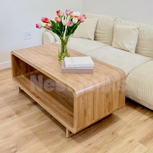 May include: A light wood coffee table with a rounded rectangular frame and an open storage shelf. On top sits a clear glass vase filled with pink and white tulips, along with a stacked book titled Henri Matisse with a light pink cover.