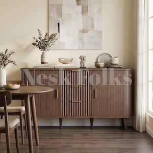 May include: A dark brown wooden sideboard featuring vertical fluted detailing on the front, three central drawers with slim gold handles, and two side cabinets. The sideboard is topped with a vase of eucalyptus, a bowl, and ceramic dishes.