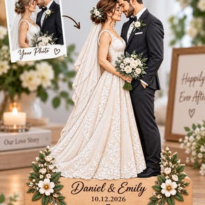 Könnte beinhalten: Ein Tortenaufsatz für Hochzeiten mit einer Braut im weißen Spitzenkleid und einem Bräutigam im schwarzen Anzug, stehend auf einem rechteckigen Holzsockel mit der Gravur Daniel & Emily, dem Datum 10.12.2026 und weißen Blumenverzierungen.