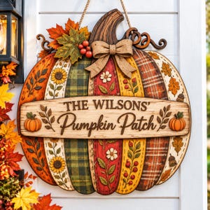Könnte beinhalten: Ein hölzernes Türschild in Kürbisform mit einem horizontalen Schild, das den Text THE WILSONS' Pumpkin Patch trägt. Das Design umfasst bemalte Abschnitte mit Karomustern und Blumenmotiven, eine Juteschleife, Herbstblätter, Beeren und kleine Kürbisse.