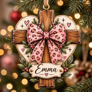 Könnte beinhalten: Ein weihnachtlicher Schmuckanhänger in Form eines Holzkreuzes, das über einem Herz liegt. Er ist mit einer großen rosa Schleife mit Leopardenmuster, grünen Blättern mit roten Beeren und einem Schild mit dem Namen Emma in schwarzer Schrift verziert.