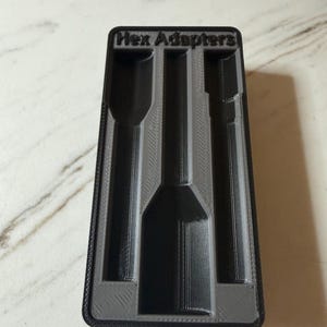 Puede incluir: Una bandeja de almacenamiento rectangular negra impresa en 3D con un interior gris que presenta tres canales verticales empotrados para herramientas. El borde superior tiene el texto Hex Adapters en relieve negro sobre fondo gris.