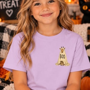 May include: A light purple short-sleeved t-shirt featuring a graphic on the left chest area. The graphic depicts a giraffe wearing a white ghost costume with the word BOO printed in black capital letters on the front of the ghost sheet.