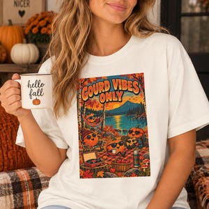 May include: A white t-shirt featuring a graphic print with the text GOURD VIBES ONLY. The illustration depicts anthropomorphic pumpkins relaxing by a lake, with one in a hammock, one playing guitar, and one sitting on a plaid blanket with a record player.