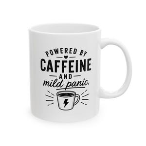 Op de afbeelding: Een witte keramische mok met zwarte tekst die luidt POWERED BY CAFFEINE AND mild panic. Een klein hartpictogram staat boven het woord CAFFEINE en een afbeelding van een koffiekopje met een bliksemschicht staat gecentreerd onder de tekst.