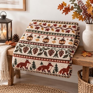 May include: A cream-colored crochet blanket featuring a repeating autumn-themed pattern. The design includes rows of orange and brown foxes, green pine trees, brown pinecones, and hanging lanterns, all accented by borders of fall leaves.