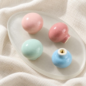 May include: Four round ceramic cabinet knobs in pastel colours including light pink, dusty rose, mint green, and sky blue. Each knob features a smooth, glossy finish and a central metal threaded insert on the underside for mounting purposes.